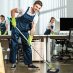 young cleaner looking at camera while washing floor with mop in modern office